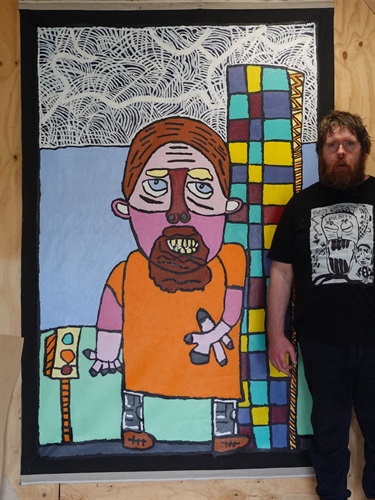 Artist Matthew Clarke standing beside one of his large-scale artworks selected for the Creative Moyne temporary public art program. The colourful self-portrait features a pink-faced figure in an orange shirt standing beside a tall patterned building, painted in Matthew's distinctive expressive style.