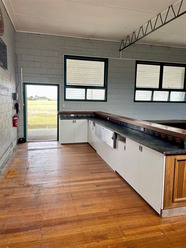 Interior of Killarney Pavilion showing bar service area with counter, cabinetry, timber floors, and doorway opening to the sports field outside.