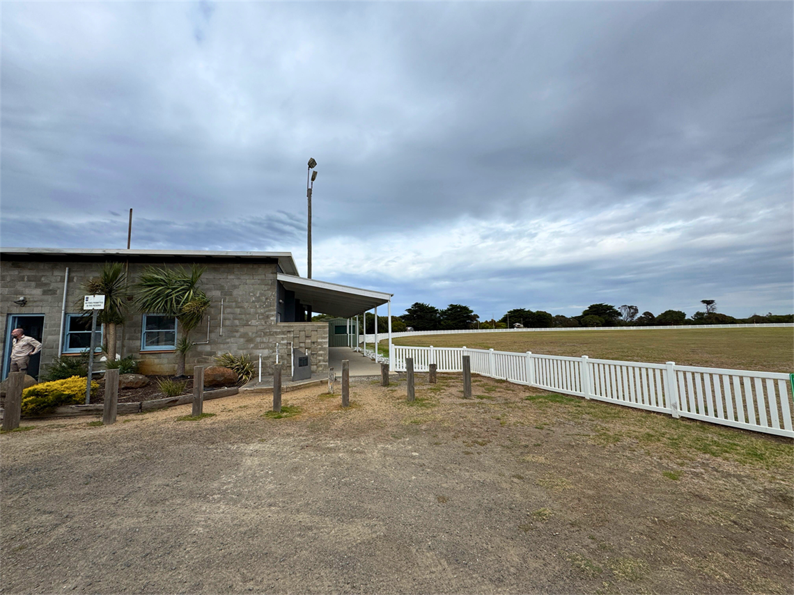 Exterior of Killarney Pavilion, a stone community building beside an open sports field, with a covered walkway and white boundary fence.