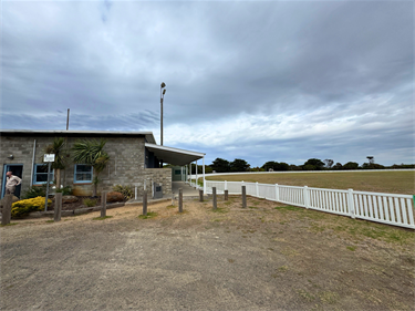 Exterior of Killarney Pavilion, a stone community building beside an open sports field, with a covered walkway and white boundary fence.