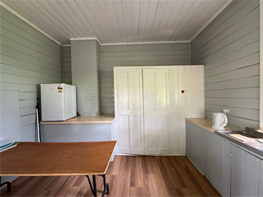 Interior of Montgomery Pavilion showing a light-filled timber room with grey wall panelling, wooden floor, kitchenette with sink and kettle, fridge, storage cupboard and table workspace.