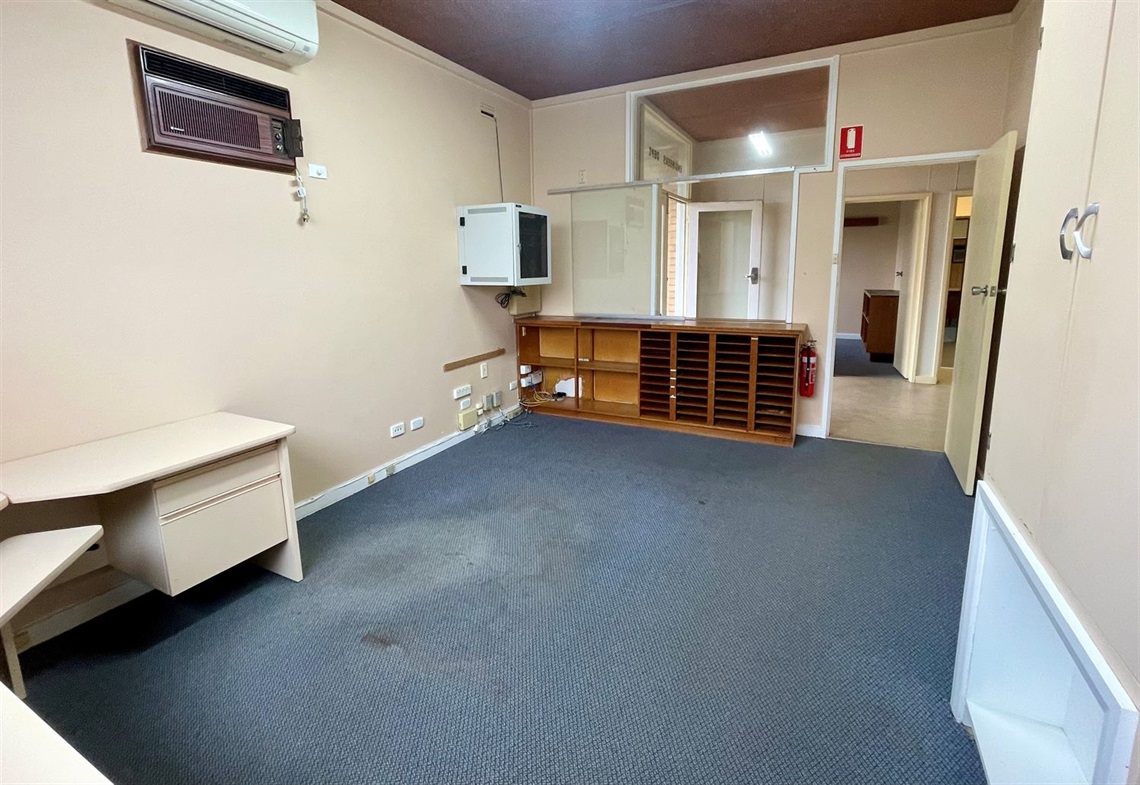 Studio Two at Macarthur Depot showing a larger carpeted workspace with desk, wall shelving, air-conditioning unit, and doorway connecting to other rooms.