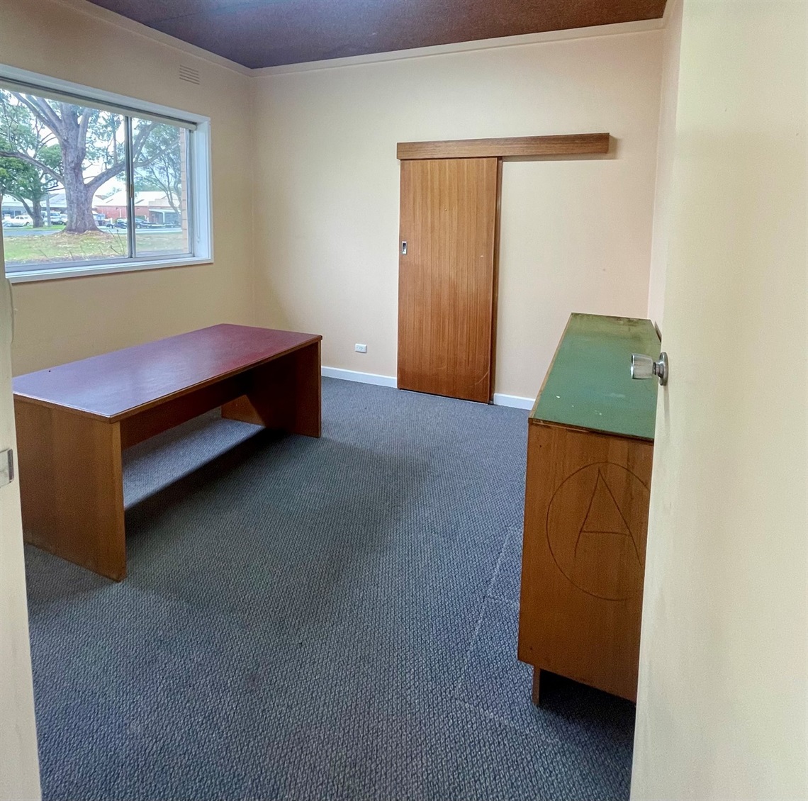Studio One at Macarthur Depot showing a small carpeted workspace with a desk, storage unit and window overlooking trees, with a sliding door cupboard.