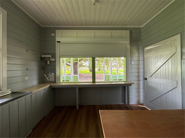 Interior of Montgomery Pavilion showing timber-lined walls, kitchenette bench with sink, wooden floors, and a large service window opening to views of the surrounding park.